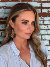 Woman with long blonde hair and earrings standing in front of a brick wall.