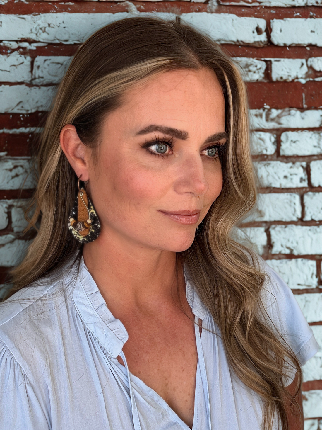 Woman with long blonde hair and earrings standing in front of a brick wall.